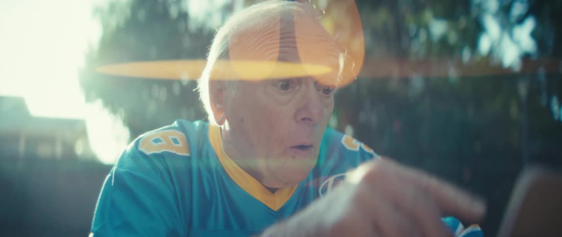 An elderly man stands outdoors under the setting sun watching a sports game on his phone and bursting with excitement