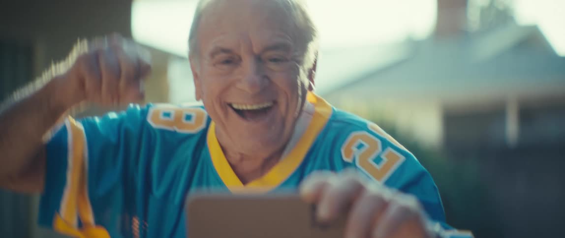A sundrenched closeup shows an older man wearing a blue and yellow sports jersey that reads blaze he is holding a smartphone out in front of him cheering with an ecstatic openmouthed smile suggesting a joyous reaction to a mom