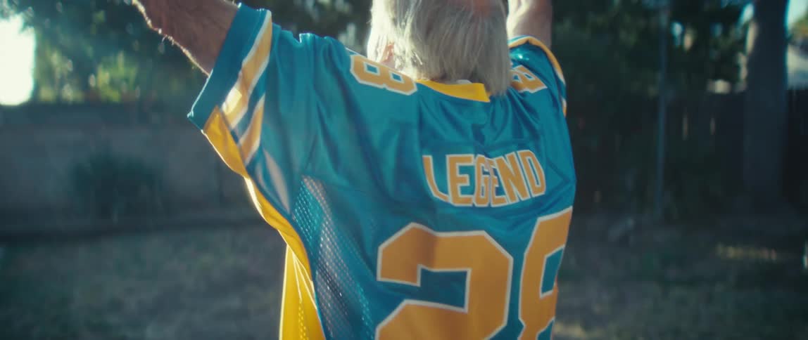A male with gray hair wears a blue and yellow sports jersey and raises his arms up while celebrating outdoors