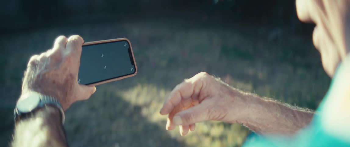 An elderly man stands outdoors under the setting sun watching a sports game on his phone