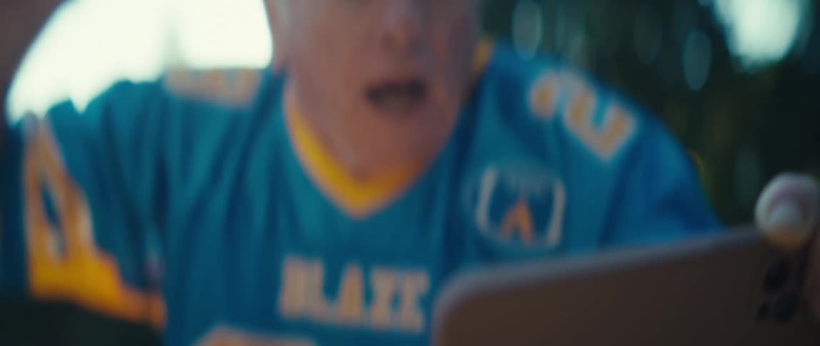 A sundrenched closeup shows an older man wearing a blue and yellow sports jersey with a shocked openmouthed expression as he looks at an object below the frame he lifts his hands and his face transforms into a wide ecstatic smile as