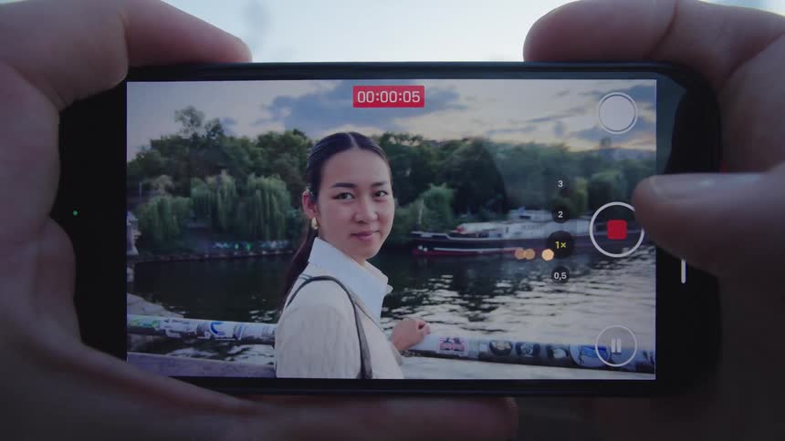 A person records a video of a young woman standing on the side of a bridge at midday holding the handrail and striking a pose