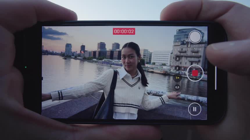Pair of hands holding a smartphone and photographing a young woman by a lake with buildings in the background