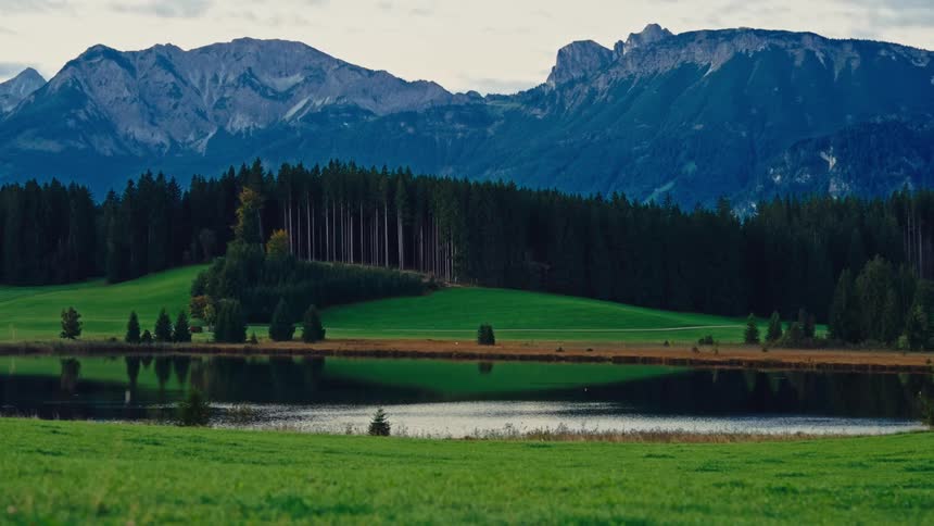 A calm lake sits below green fields and tall mountains