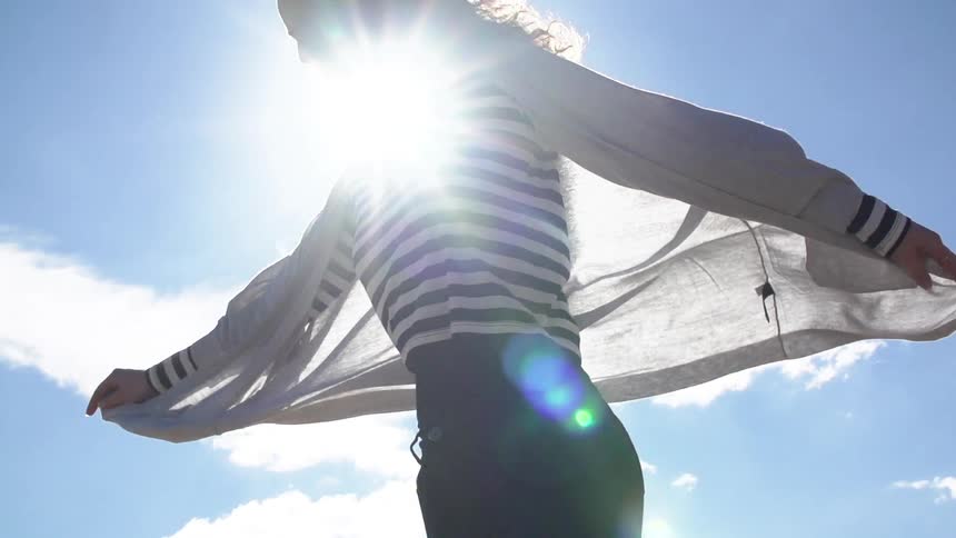 A woman holding her jacket out so the breeze catches it while she spins around with the sun glaring behind her