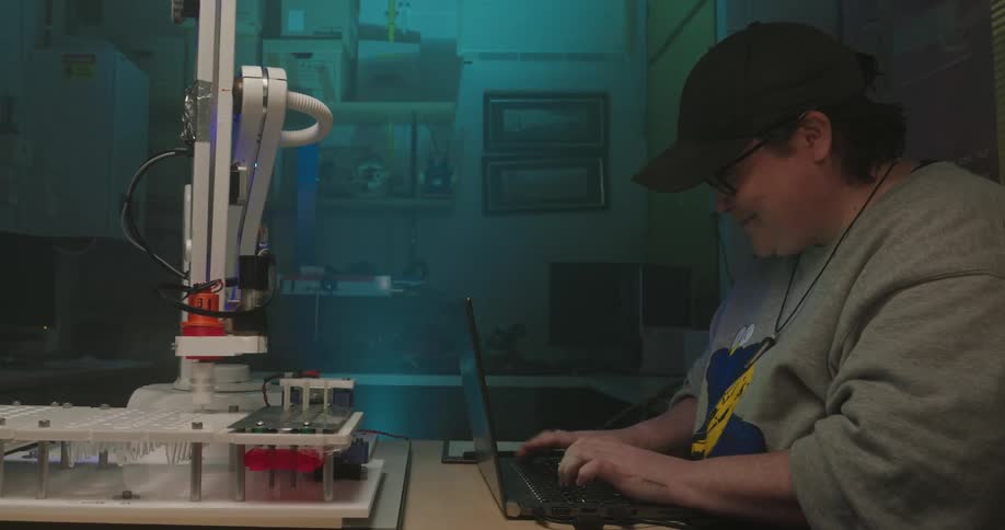 Person typing on a laptop next to a robotic arm in a laboratory environment