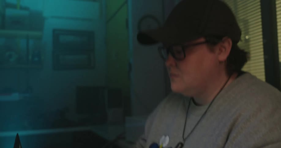 A young man wearing a cap and glasses sits at a desk and works on a laptop while a small white and orange robotic arm performs a precise operation in the foreground