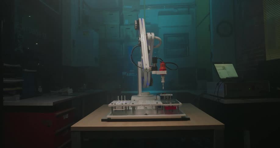 A small white and red robotic arm stands on a wooden table in the center of a darkened room and performs vertical and rotational movements over a rack of sample containers