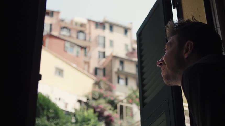 Close up of a young man standing contemplatively at a window indoors during daytime in a suburban city