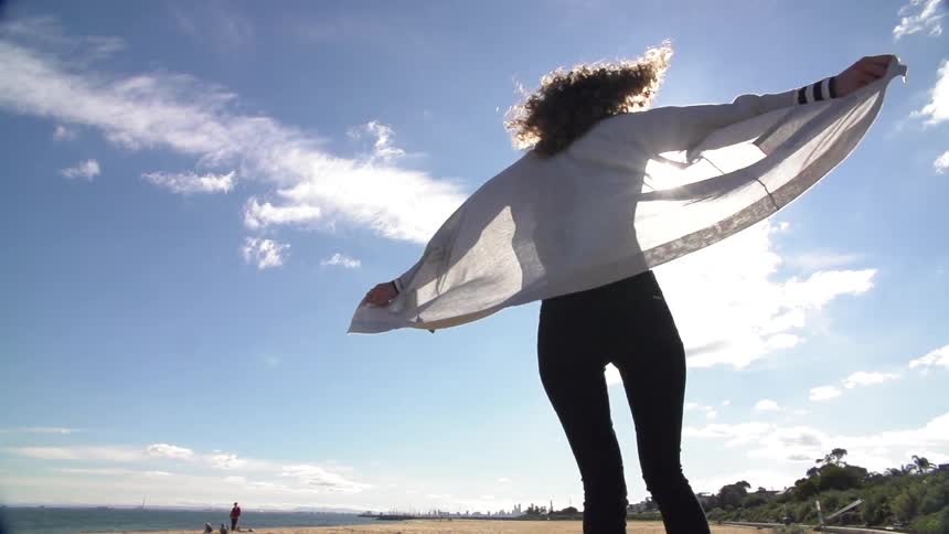 A woman on a beach holding her jacket out so the breeze catches it while she spins around with the sun glaring behind her
