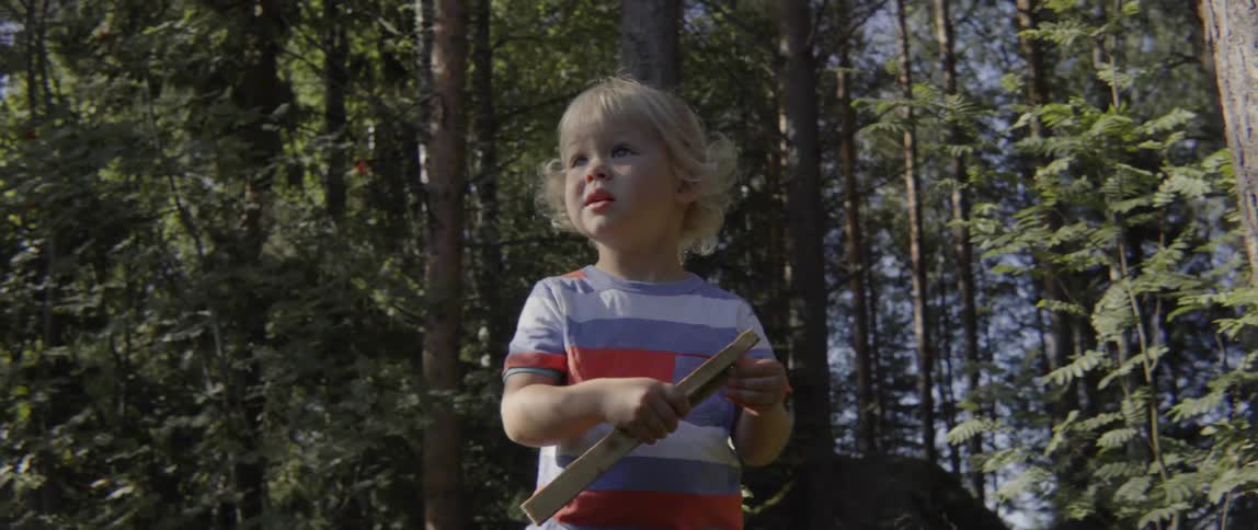 Young toddler boy looking around curiously while holding a stick in a sunlit forest.