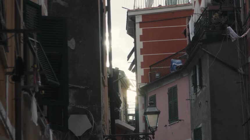 A narrow space between multistory buildings with closed shutters and balconies while bright light shines from the sky through the top of the passage