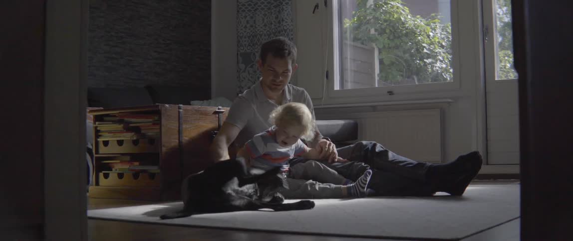 Young father and toddler boy petting a large black dog while sitting together inside.