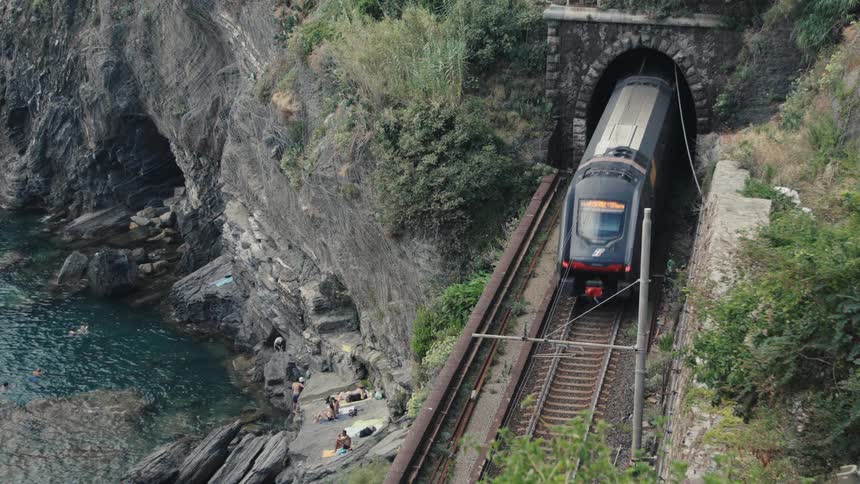 A passenger train emerges from a stone tunnel entrance built into a steep cliffside traveling along railroad tracks above rocky coastal water