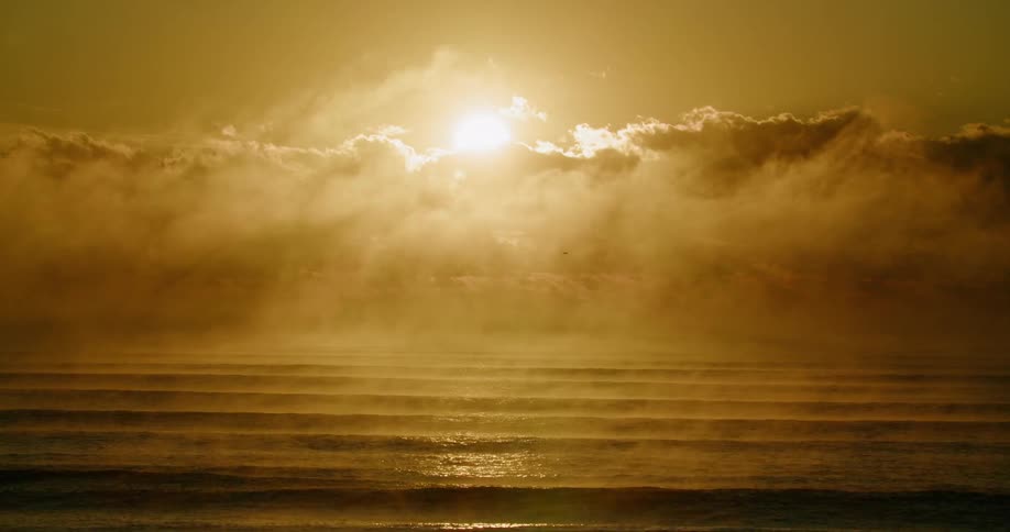 A large sun shines intensely through horizontal cloud cover and thick mist illuminating multiple ocean waves that roll toward a dark foreground shore
