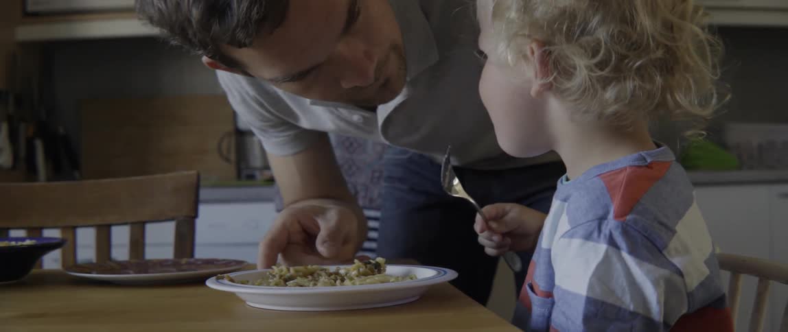 A young boy eats pasta while his dad leans over him