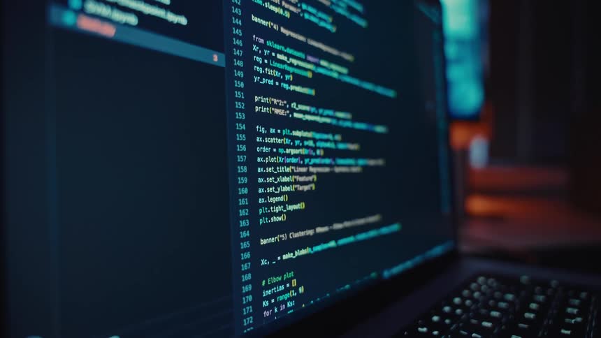 Laptop displays computer code while sitting on a table in a room containing other computers