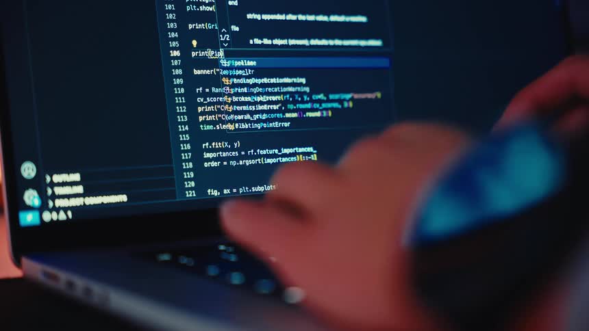 A highangle closeup shot of a laptop screen displays lines of code with bright yellow cyan and red syntax highlighting on a dark background the scene shows a hand typing on the illuminated keyboard in the foreground partially obscuring the screen