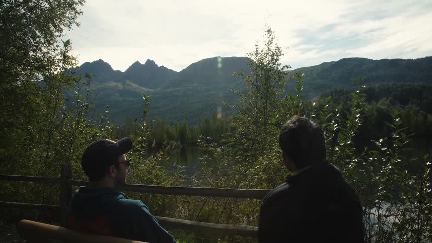 Two men sitting on a bench talking and admiring a beautiful landscape with a large lake and big mountains in the background