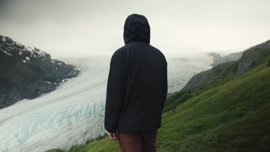 A person stands on hillside overlooking glacier under misty sky