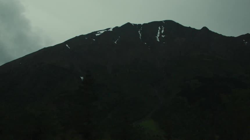 Light amount of snow sits on the summit of a mountain towering over a conifer forest on an overcast winter day