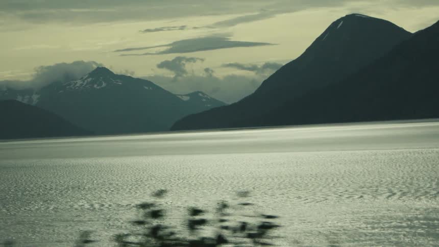 A closeup shot captures the rippling surface of a large dark body of water possibly a lake or fjord surrounded by distant mistshrouded mountains the water reflects the subdued light giving it a bright metallic sheen while the foreground shows da