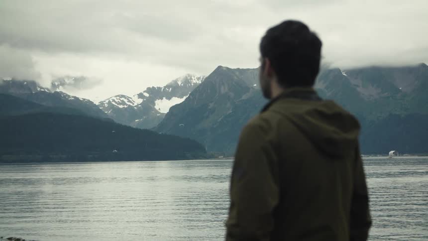 Man observing a lake surrounded by snowcapped mountains under a cloudy sky