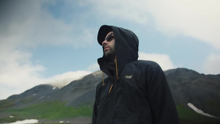 Man in hoodie stands in green mountains under cloudy sky