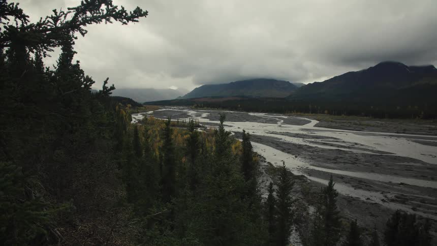 Winding river cuts through dense forest under a cloudy sky between mountains