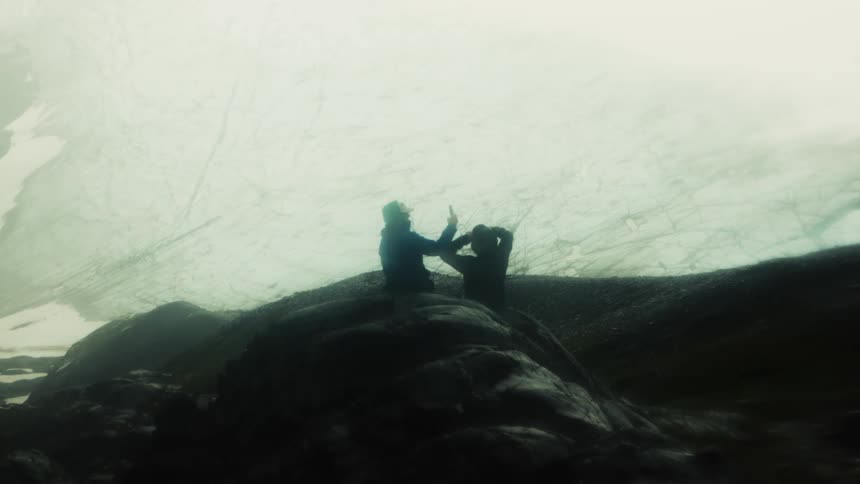 Two silhouetted people sit on rocks photographing a massive hazy ice wall