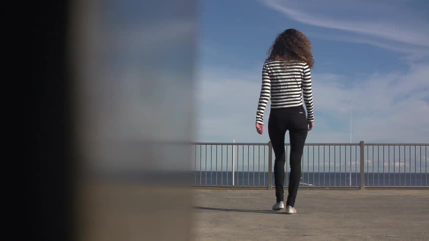A woman wearing a black and white striped sweater walking towards a railing next to the ocean
