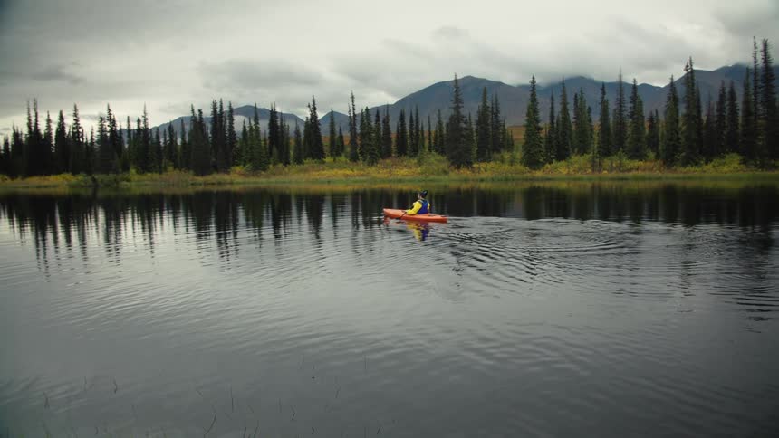 Person paddling an orange kayak on a calm lake surrounded by pine trees and mountains