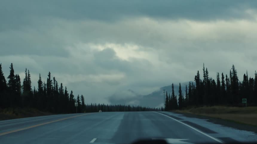 A mediumlong shot shows a straight empty wet asphalt road stretching into the distance under a dark heavily overcast sky the road is bordered on both sides by dark tall evergreen trees with a misty mountain range faintly visible in the far backgrou