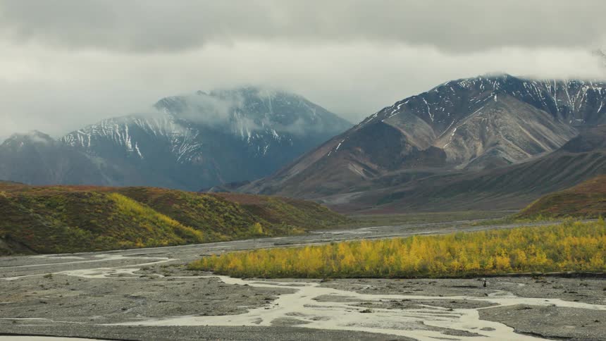 The video captures a wide landscape of majestic cloudshrouded snowdusted mountains flanking a wide riverbed with braided streams and autumn foliage the camera slowly zooms out emphasizing the grand scale of the rugged expansive valley