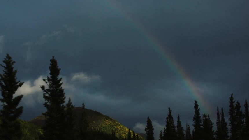 A rainbow stretches over forested mountains under dark clouds