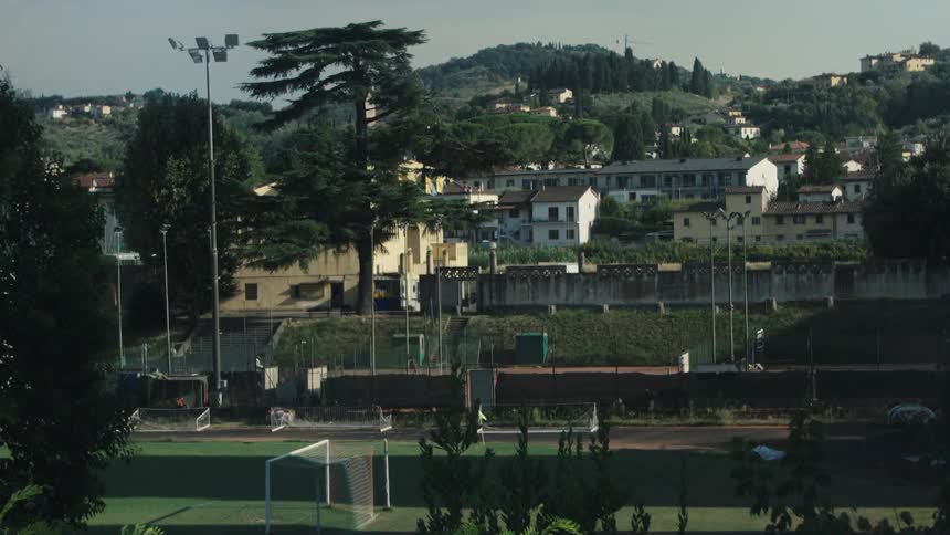 Soccer field with goalposts surrounded by fences and residential buildings in front of wooded hills