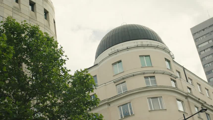 Building with dark dome surrounded by modern structures and green tree under cloudy sky