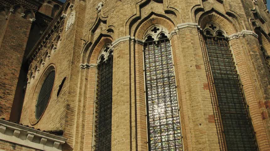 A vertical lowangle video shows the ornate brick exterior of a gothic cathedral with three tall arched windows featuring geometric stained glass and stone tracery with sunlight glancing off the textured facade