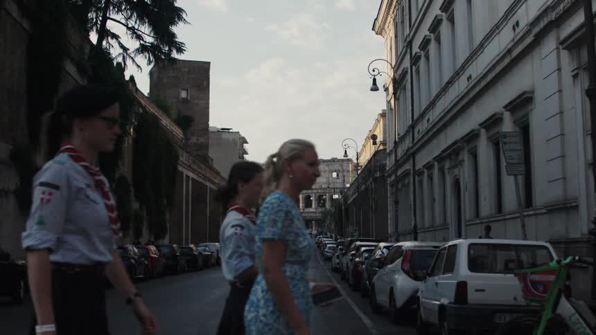 The video shows a street scene in a city with the colosseum visible in the distance down the center of the roadin the foreground on the right side of the street several people walk toward the right side of the frame including a woman in a light bl