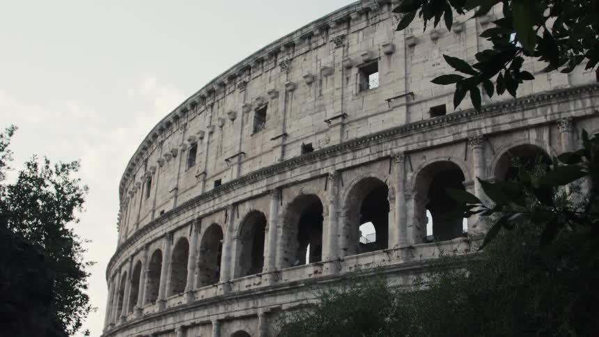 A lowangle zoomin video captures the massive curved and decayed stone exterior of the roman colosseum with its rows of arched openings partially framed by dark foliage against a bright overcast sky