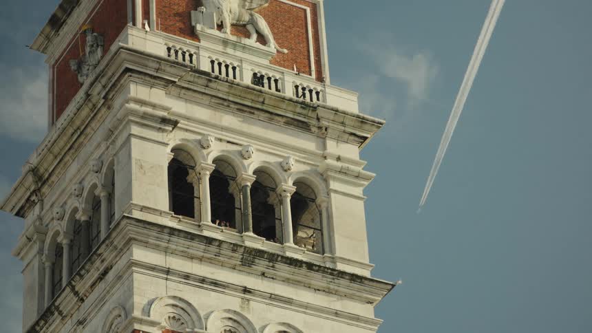 The video is an extreme closeup of the upper section of a white stone bell tower or campanile likely st marks campanile in venice italythe belfry level features a row of tall arched windows and above it a red brick section displays a large