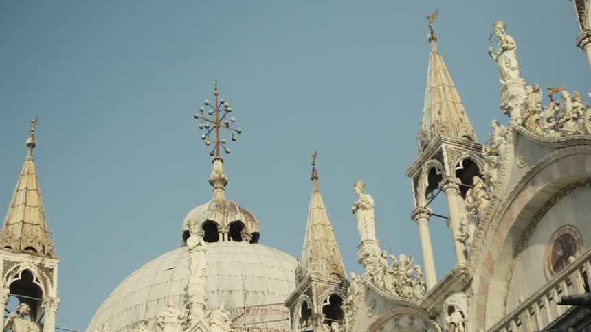 Sunlit domes and ornate spires of st marks basilica in venice rise against a clear blue sky crowded with statues and delicate carvings