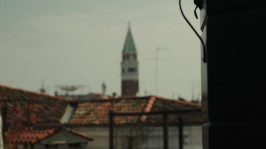 A cityscape with terracotta rooftops and a distant bell tower centered in the blur in sharp foreground the dark edge of a building with a dangling cable frames the right side