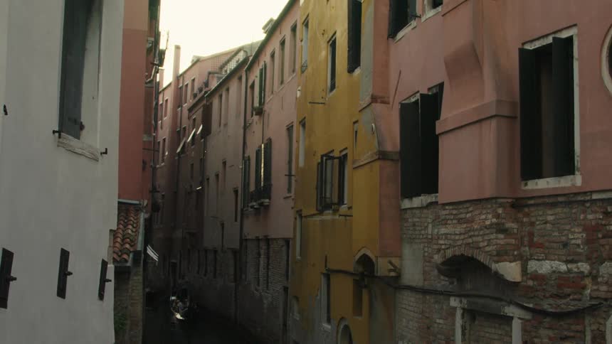 A highangle view video looks down a narrow shadowed venice canal where a dark gondola slowly glides framed by the tall colorful and weathered facades of pale pink and yellow buildings with closed black shutters