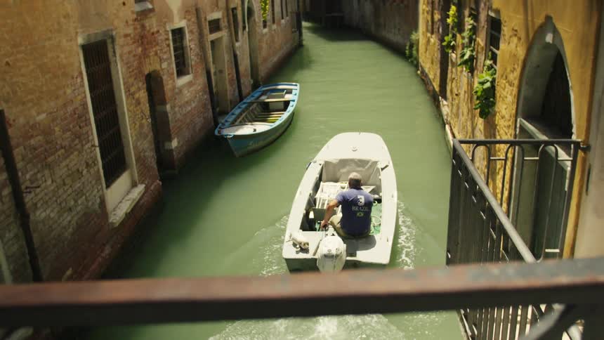 Viewed from a bridge a man in a motorboat navigates a narrow sunlit canal between the historic buildings of venice