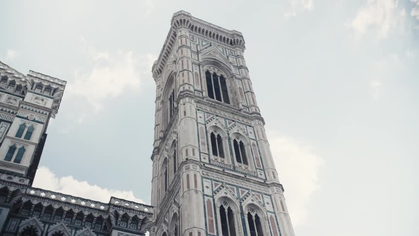 The video is an extreme closeup shot of the intricately carved white and green marble facade of the florence duomo complex moving quickly upwards the motion blurs the details of the gothic arches and columns against a bright sky