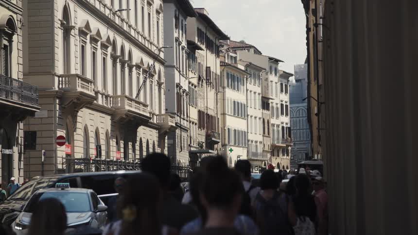 A busy city sidewalk on a sunny day crowded with people walking as cars pass along the road