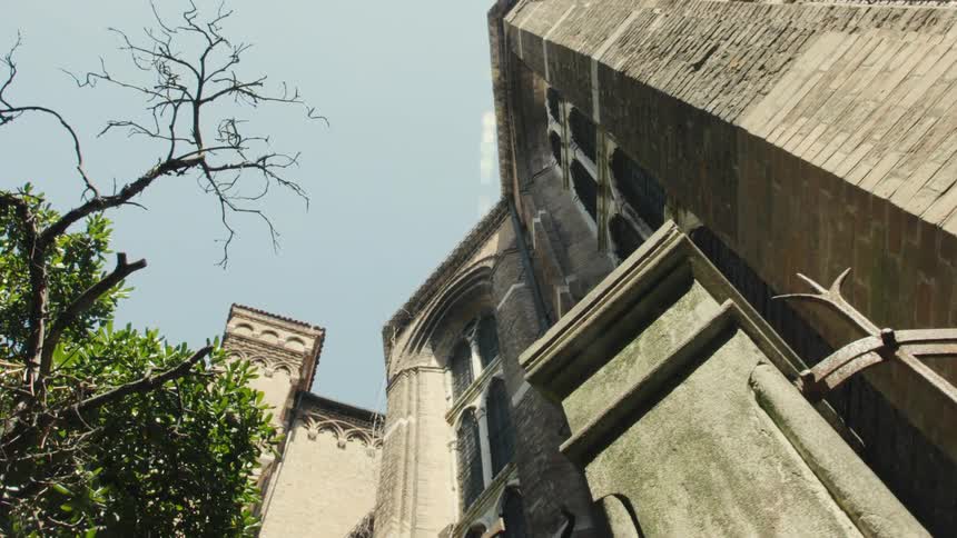 The video is an extreme lowangle upwardpanning shot of an old brick church tower and the side of a large adjacent structure the warm sunlit brick and stone architecture contrasts with the bright blue sky above and the frame includes the