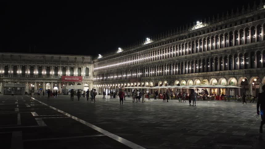The video is an establishing shot of the large open square of piazza san marco in venice italy captured at night the brightly lit multiarched architecture lining the square creates a dramatic backdrop for the small clusters of people wal