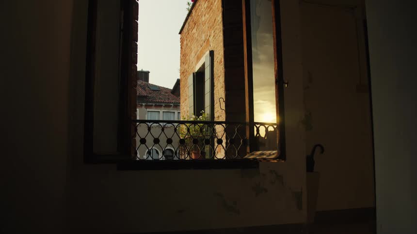 Open window reveals brick building with balcony and plants lit by sunlight